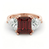 A rose gold ring with a large, deep red gemstone in the center, flanked by two smaller diamonds on either side.